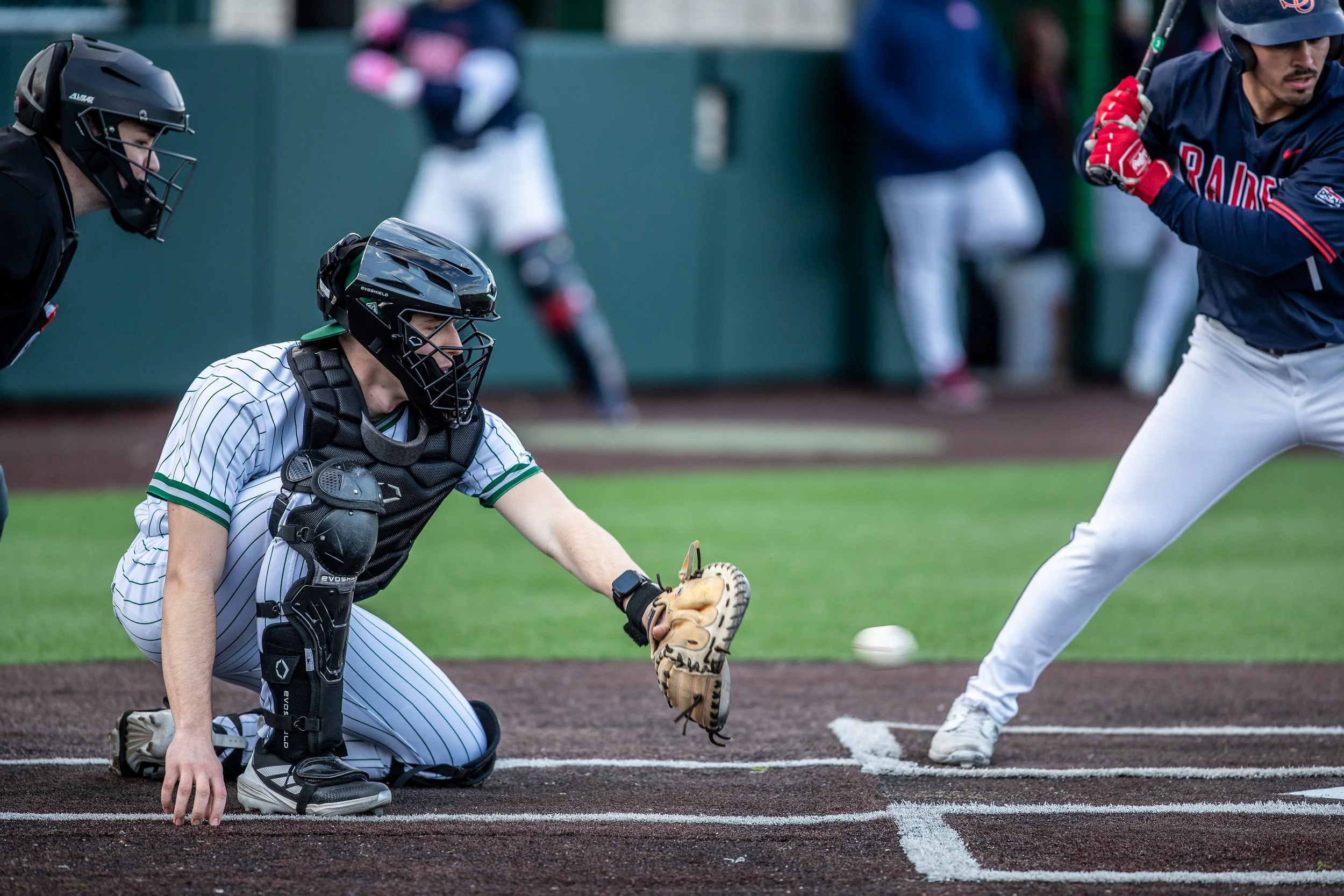 Baseball catcher kneeling behind home plate, ready to catch a pitch, with a batter swinging at a pitch.