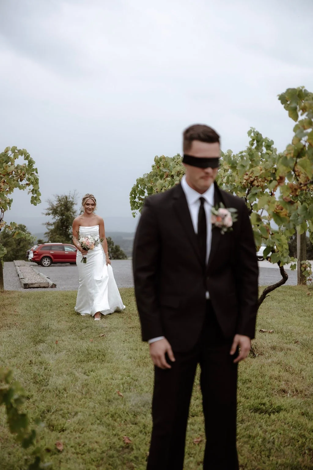 A groom in a black suit and tie, wearing a blindfold, stands in front of a bride holding a bouquet of flowers during a wedding ceremony outdoors, with trees and a cloudy sky in the background.