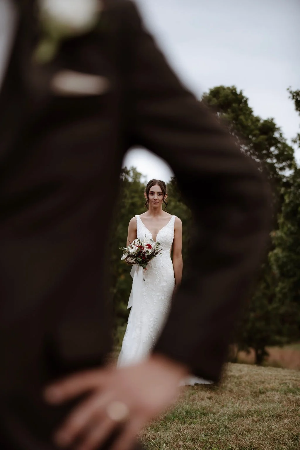 A bride in a white wedding dress holding a bouquet of flowers, standing outdoors, seen through the blurred hand of another person.