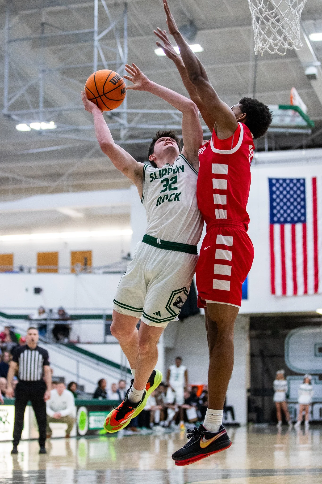 Two basketball players are jumping to contest a shot; one wearing a white jersey with green accents and the number 32, and the other wearing a red jersey with white stripes.