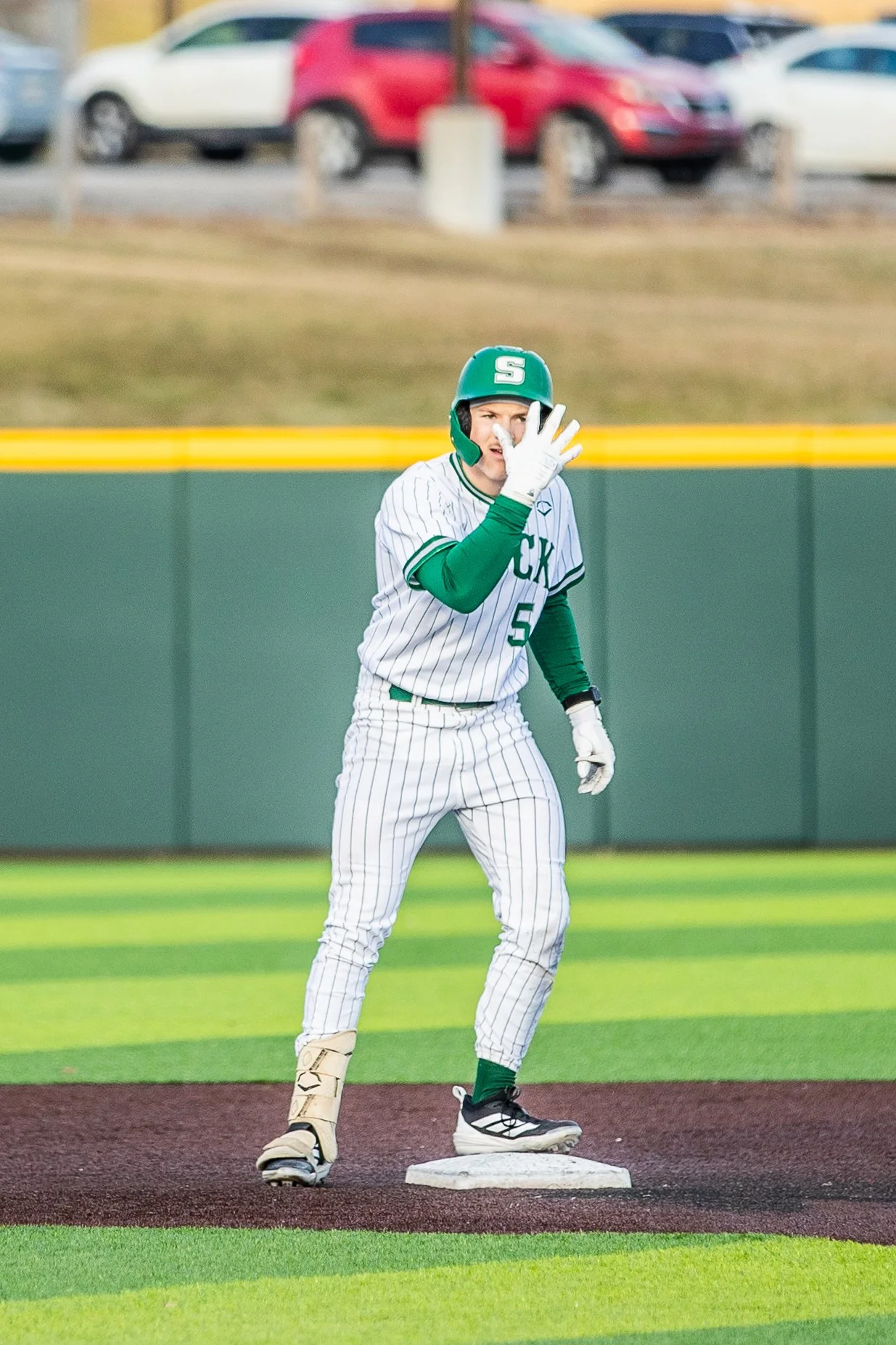 A baseball player in a pinstriped uniform with a green helmet and gloves is standing on a base, appearing to gesture or call out during a game.