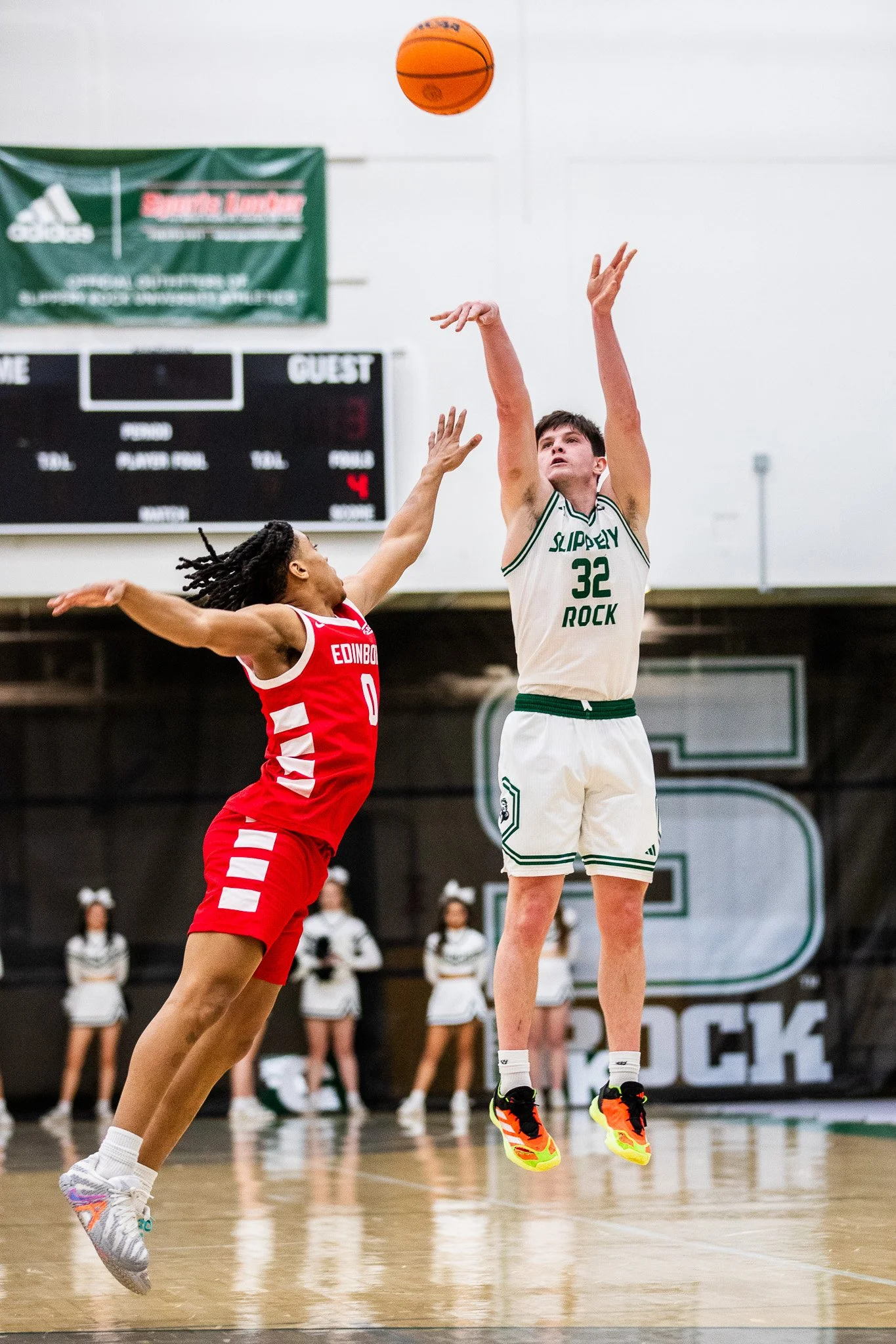 A basketball game with two players in mid-air, one in a red uniform labeled 'EDINBO' and the other in a white uniform labeled 'SLIPPERY ROCK', attempting a shot or block, with cheerleaders in the background.