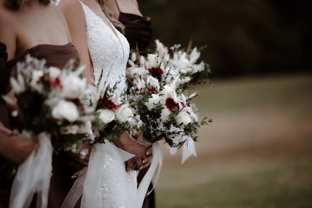Close-up of a bride in a white lace wedding dress holding a bouquet of white flowers with red accents, surrounded by bridesmaids in dark dresses holding similar bouquets.
