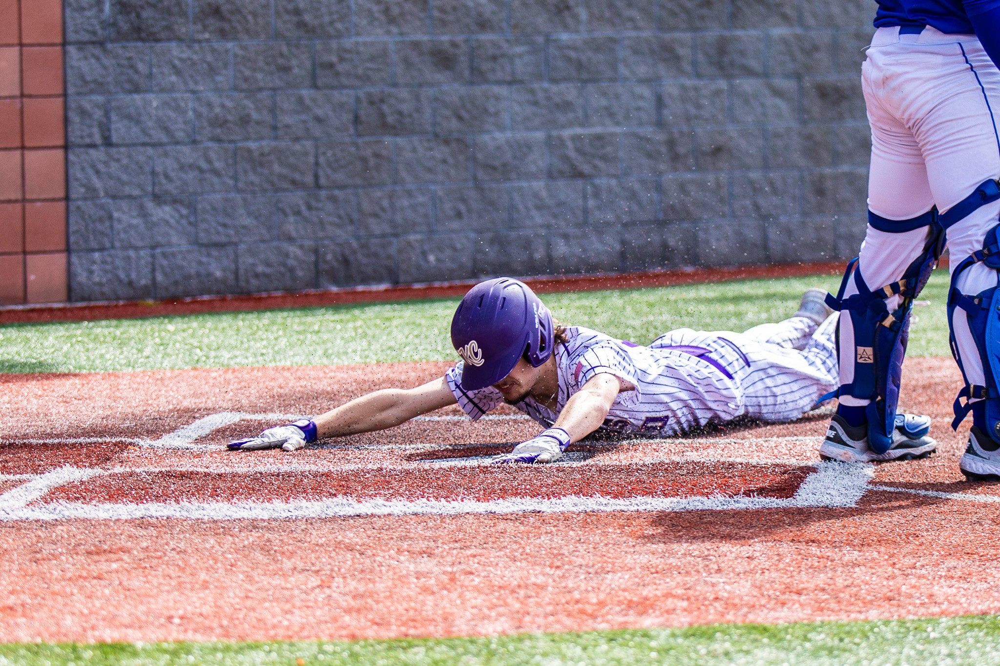 A baseball player in a purple helmet and striped uniform is sliding headfirst into a base, while an umpire or teammate stands nearby.