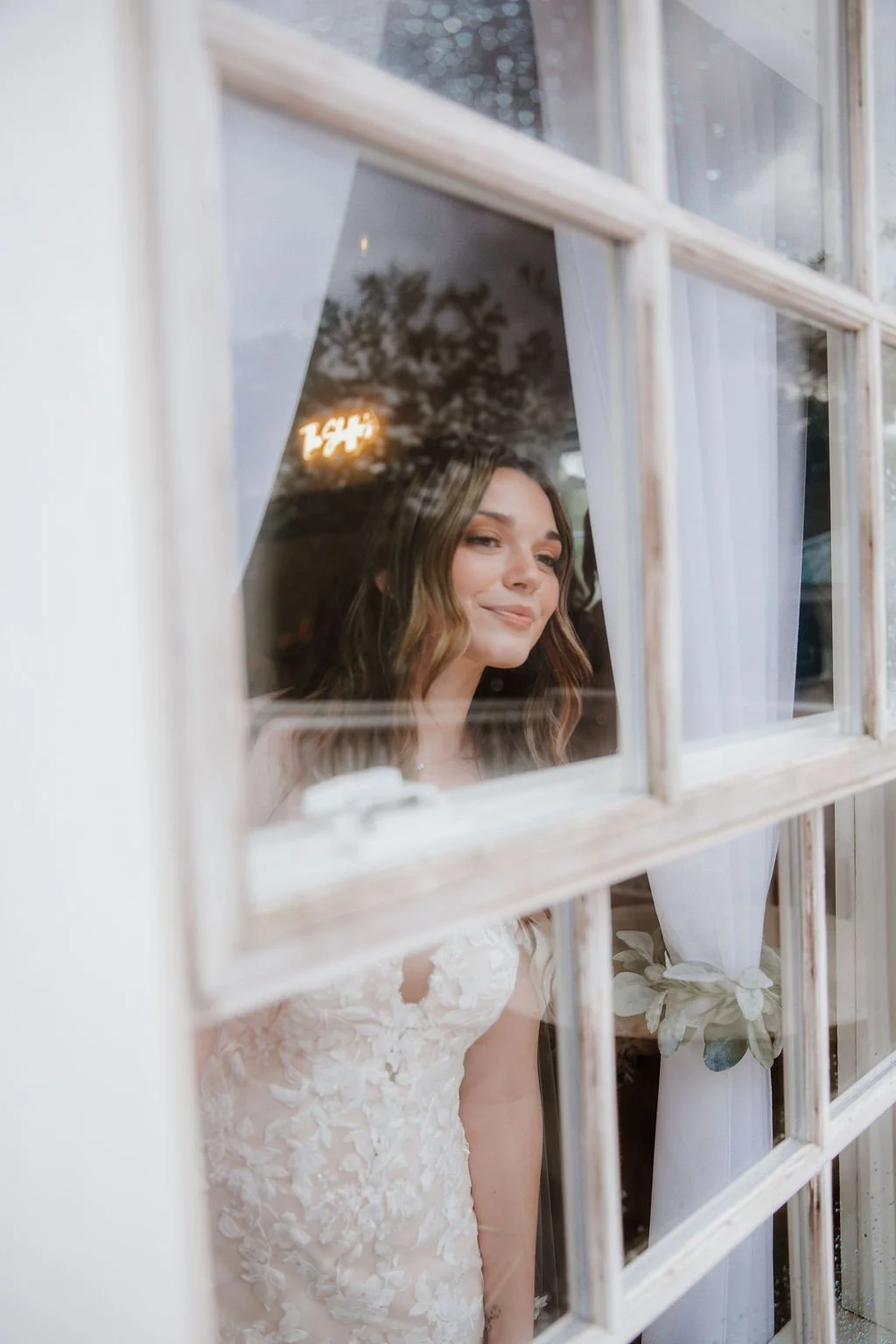 A woman in a white lace dress looking out a window with a soft smile, curtains and plants visible in the background.