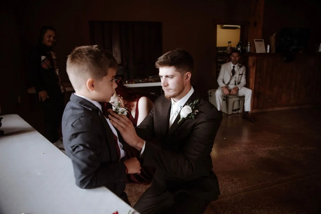 A groom kneeling in front of a young boy, possibly during a wedding ceremony, with a woman and another man in the background in a dimly lit room.