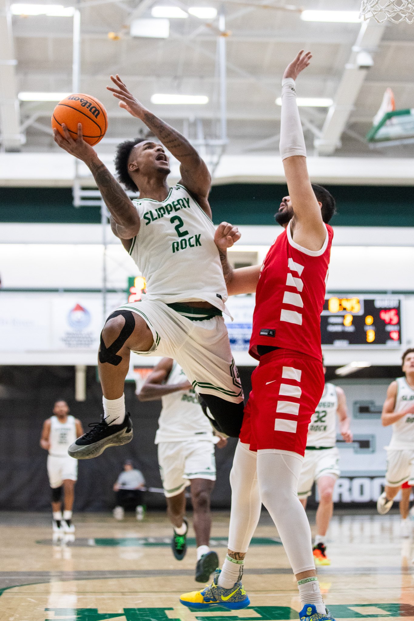 A basketball player in a white jersey jumping to shoot the ball while a player in a red jersey attempts to block the shot during a game at an indoor basketball court.