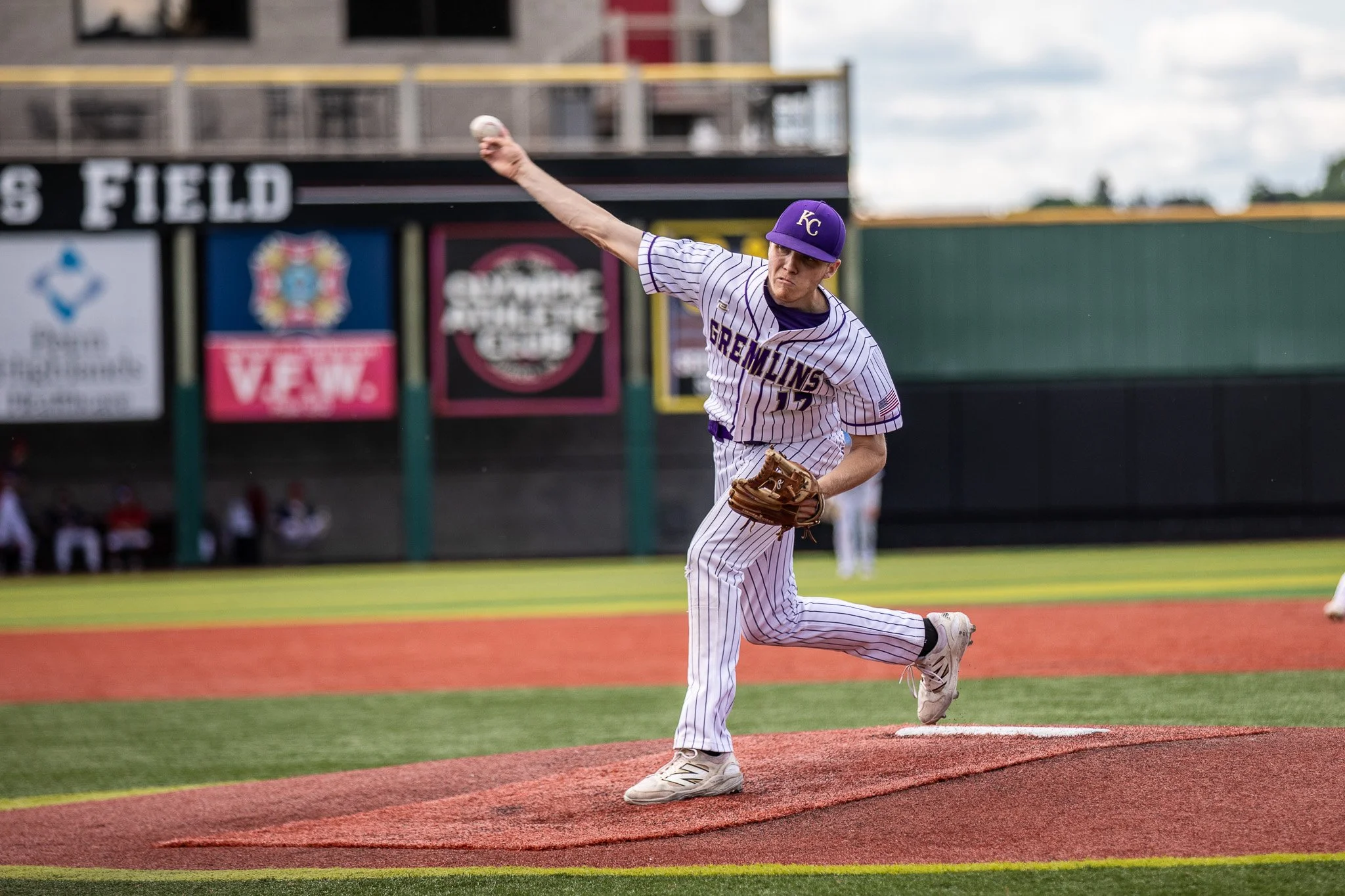 A baseball pitcher is in mid-throw on a baseball field, wearing a purple hat and a pinstriped uniform with 'GREMLINS' on the front, on a bright day.