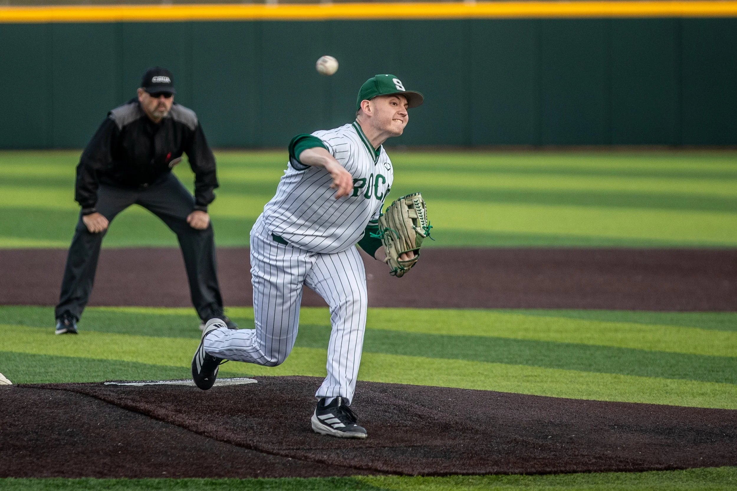 A baseball player in uniform pitching on the mound while a home plate umpire watches behind him.