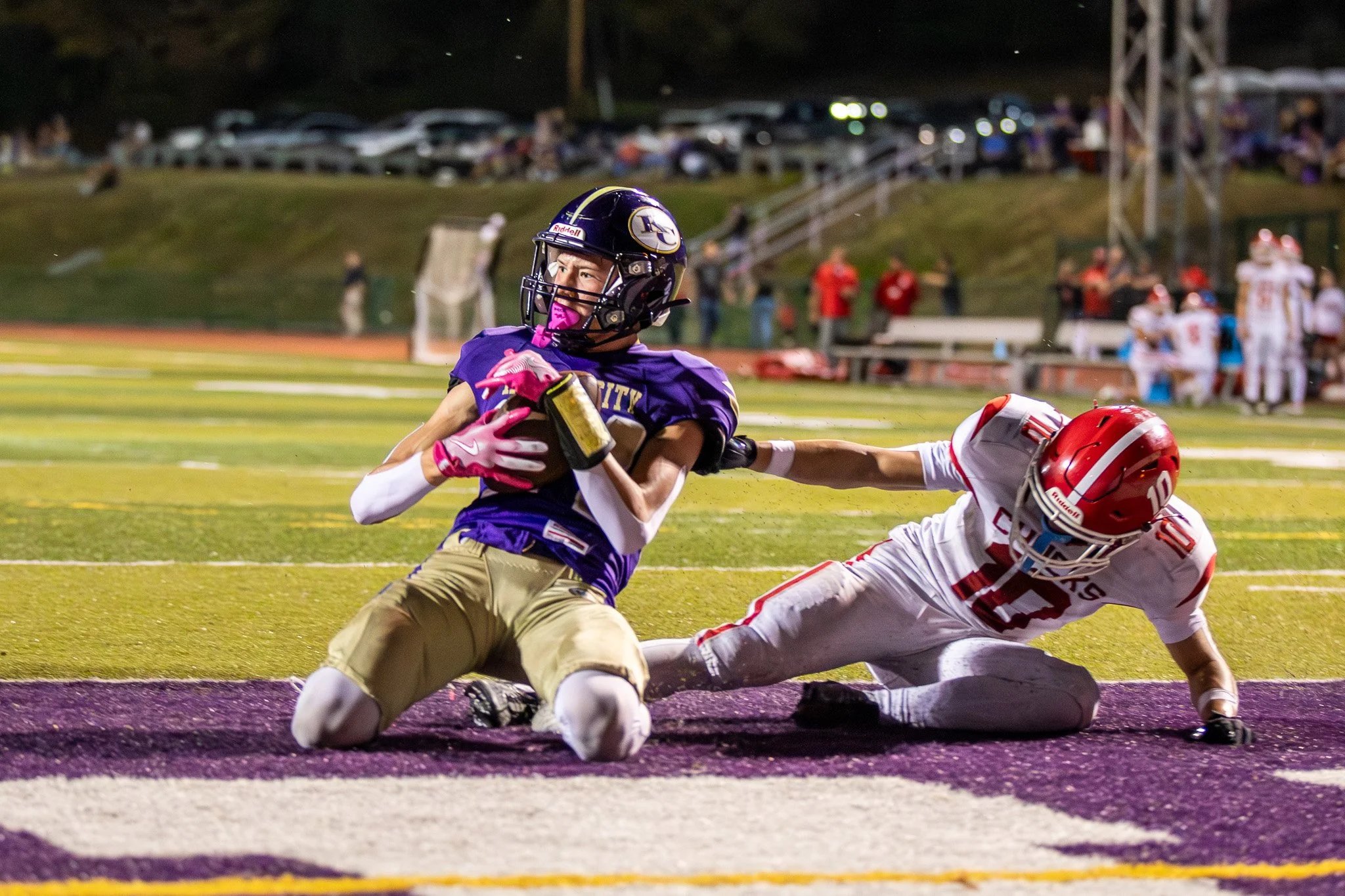 A football player in purple and gold uniform running with the ball while a player in white and red uniform is diving to make a tackle during a game at night.