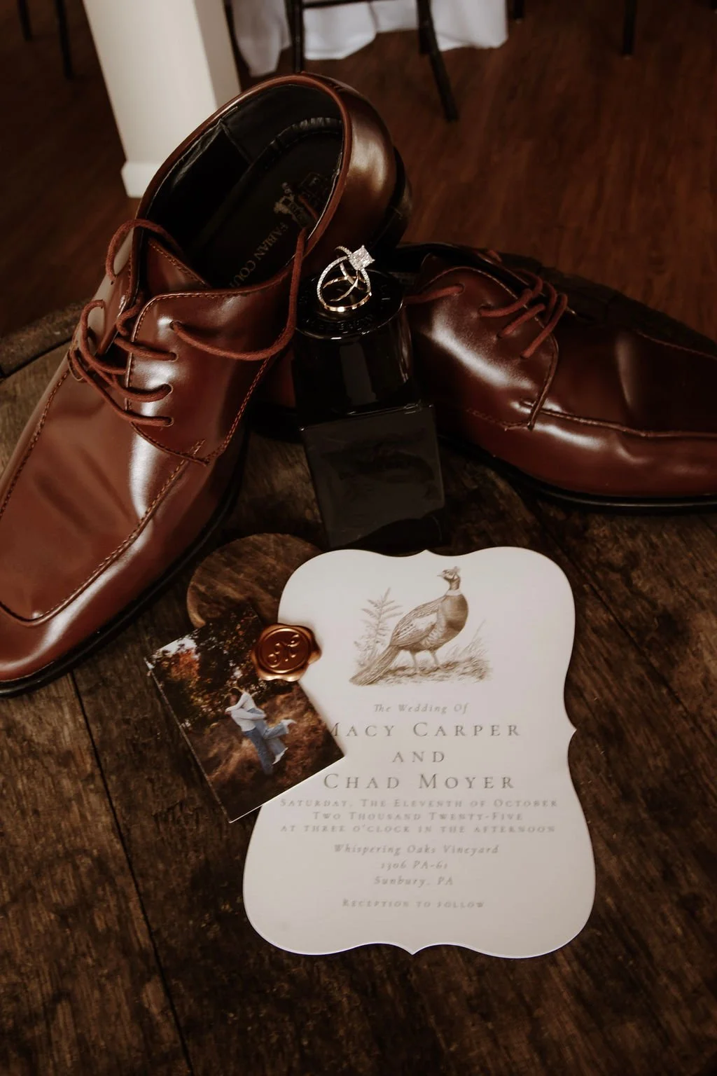 Brown leather dress shoes, a perfume bottle, wedding invitation for Macy Carper and Chad Moyer, and a wedding photo card on a wooden surface.
