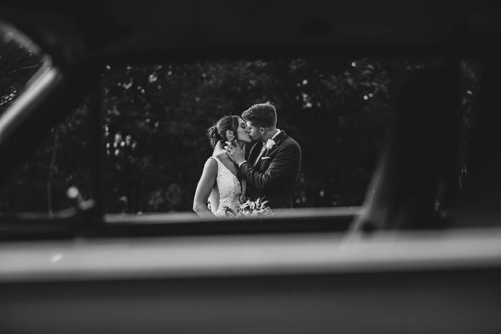 A couple in wedding attire sharing a kiss in an outdoor setting, viewed through a window.