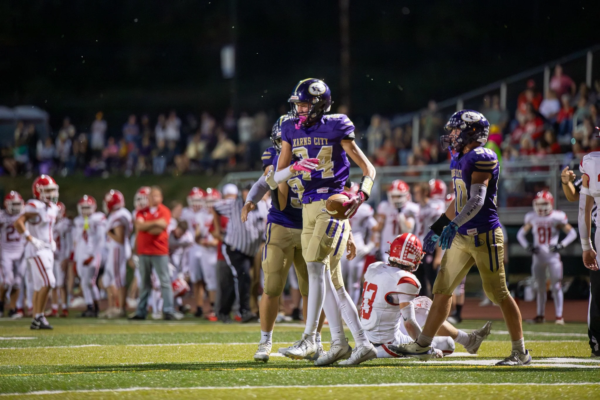A football game at night with players on the field. The team in purple uniforms, labeled 'XARRS CITY,' is celebrating as one player holding the ball stands out. The opposing team in white and red uniforms is on the ground, and players and spectators 