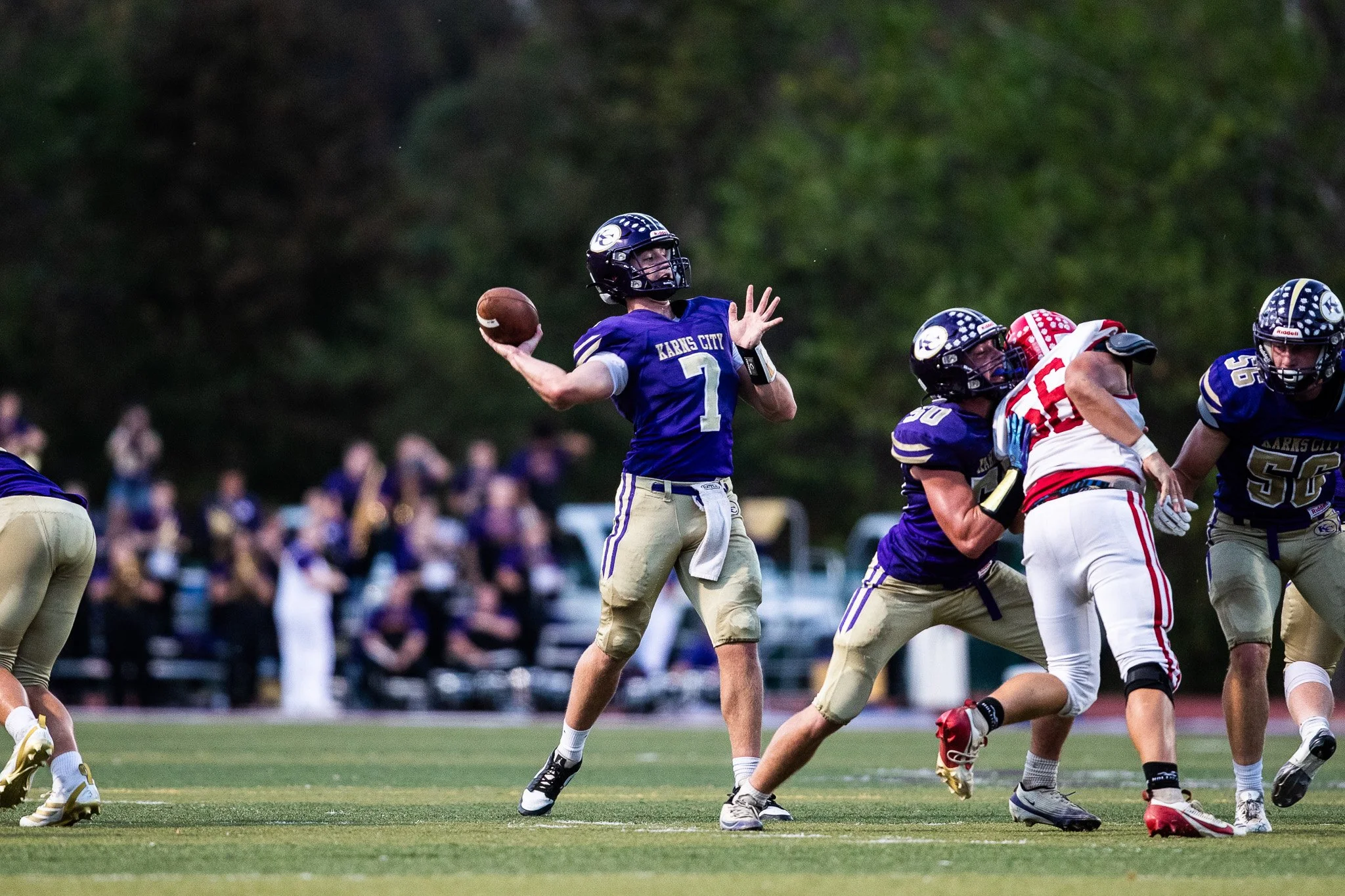 A youth football game with players in purple and white uniforms. The quarterback, wearing jersey number 7, is preparing to throw a pass while other players block opponents on the field. Spectators watch from the sidelines in the background.