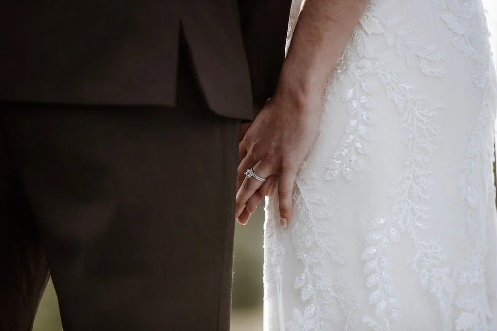 Close-up of a bride and groom holding hands, showing wedding rings, with the bride wearing a white lace wedding dress.