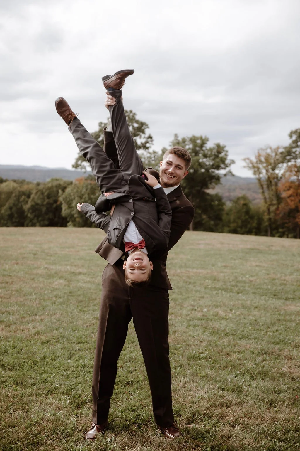 A man in a suit holding a smiling boy upside down in a grassy field with trees and cloudy sky in the background.