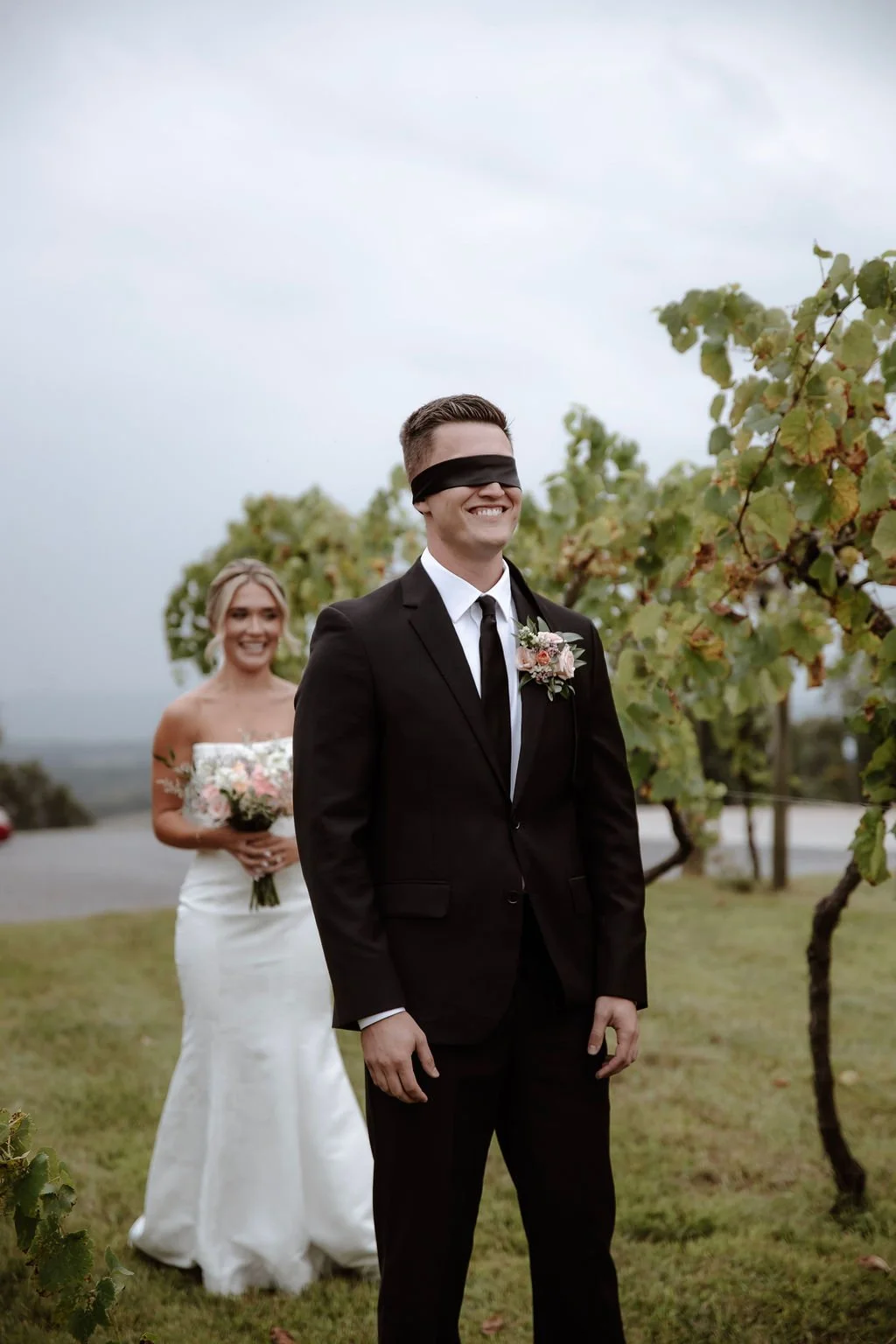 A bride and groom standing outdoors among grapevines, with the groom wearing a black suit and blindfolded, and the bride holding a bouquet of flowers behind him.