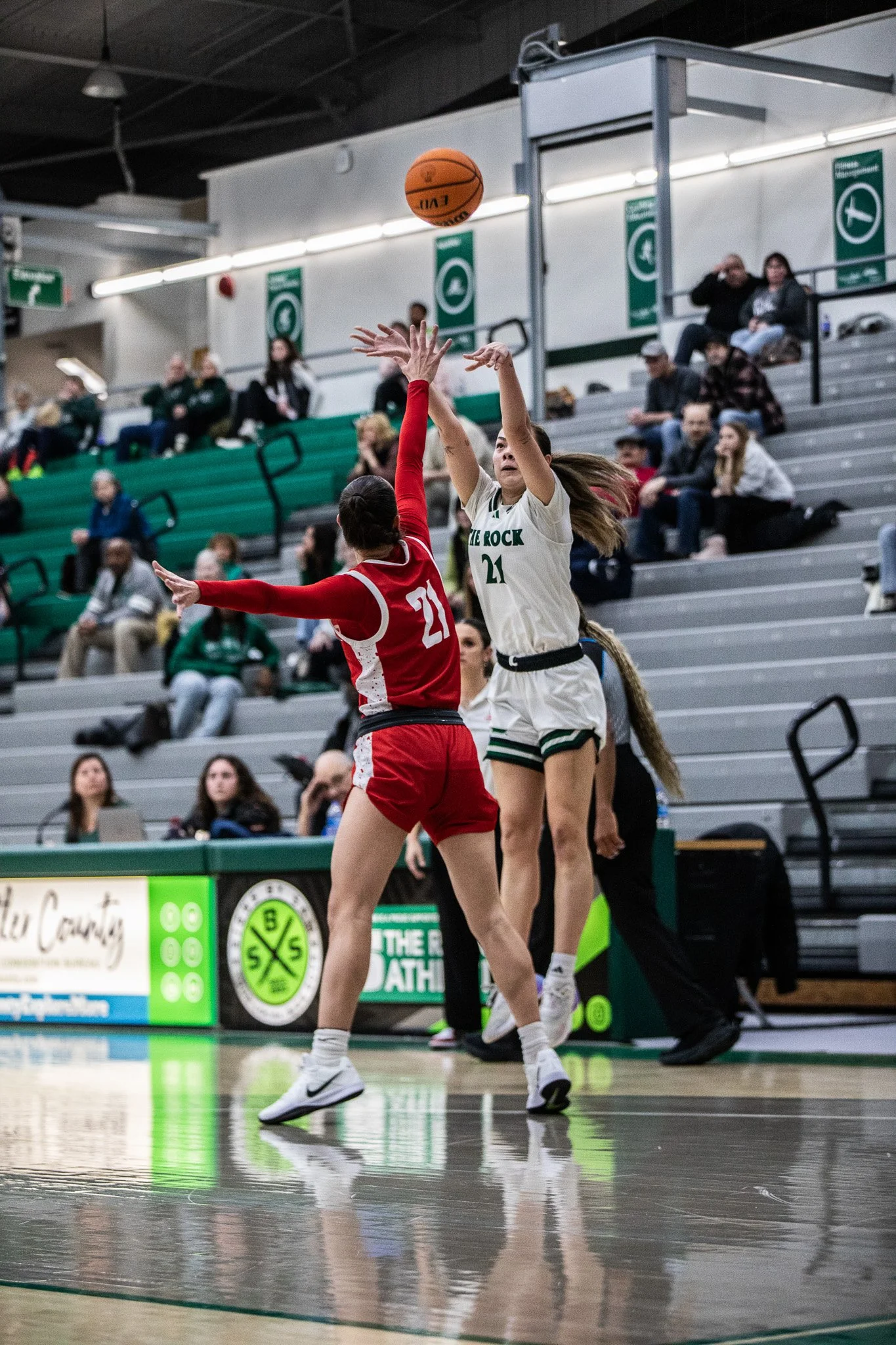 Two female basketball players, one in a red uniform and the other in a white uniform, jump to contest a basketball shot during a game in an indoor gym with spectators seated in the background.