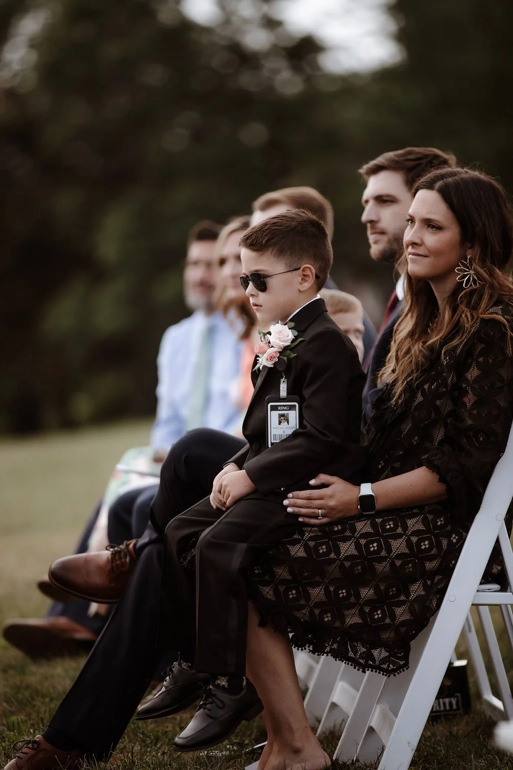 A group of people, including a young boy in sunglasses and a suit with a boutonniere, attending an outdoor event.