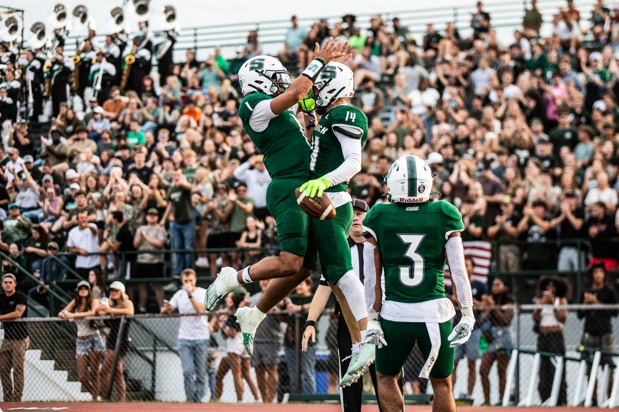 Two football players in green jerseys high-five each other on the field, with a crowd of spectators cheering in the background, while a third player stands nearby.