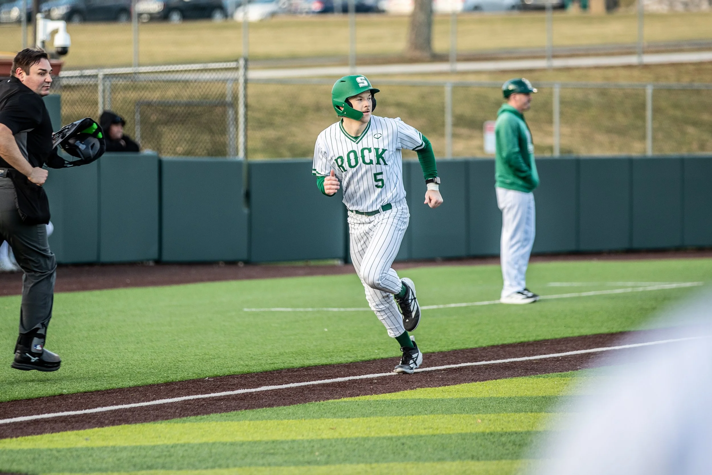 A baseball player wearing a gray and green uniform with the number 5 on it, running along the sideline during a game. There is a coach or team staff member in a green jacket and white pants in the background, and an umpire running next to the player.
