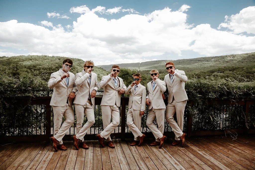 Group of six men in beige suits and sunglasses posing on a wooden deck with a railing, green trees, hills, and a partly cloudy sky in the background.