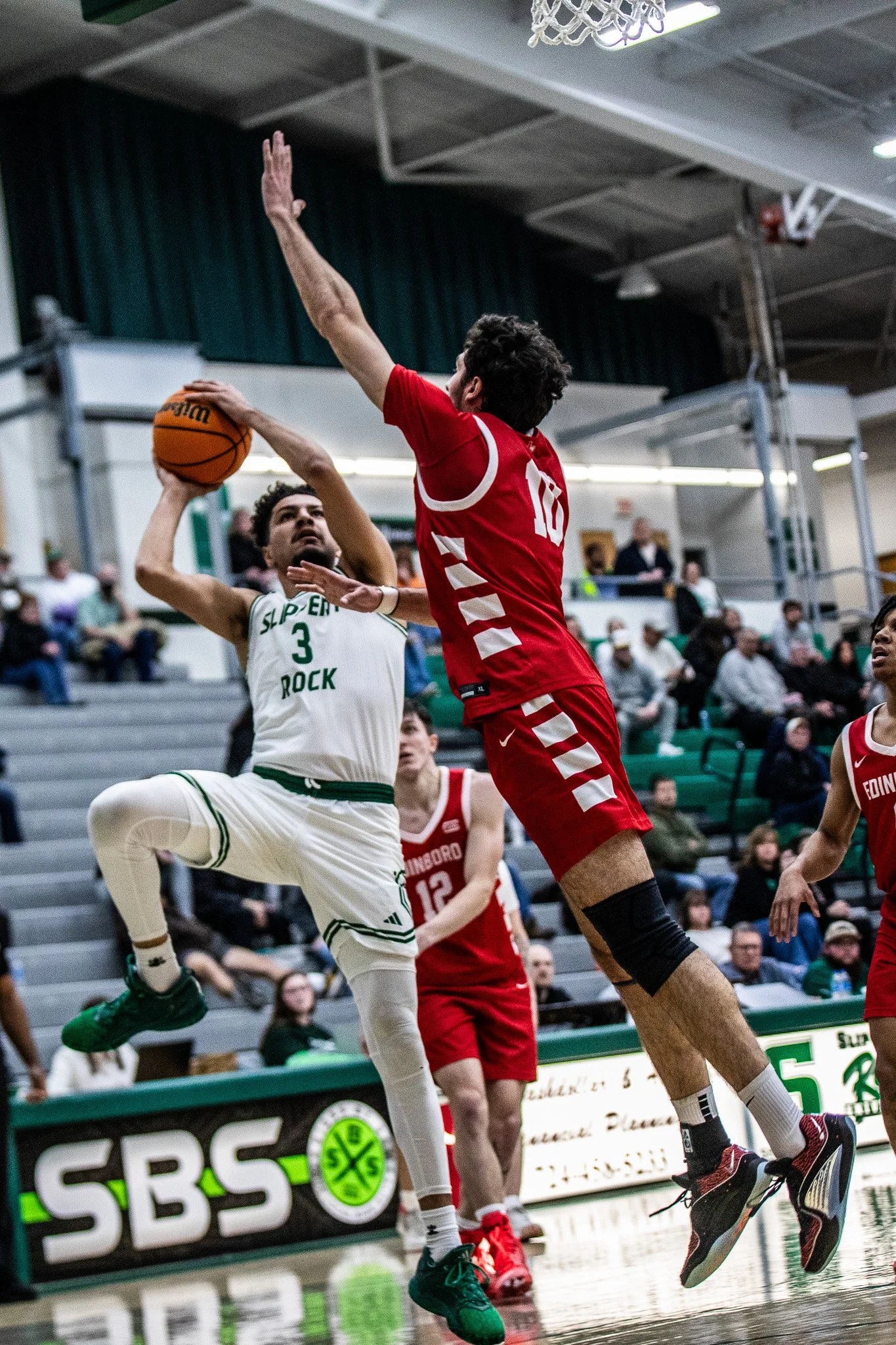 A basketball player in a white uniform with green accents jumping to shoot the ball while a player in a red uniform with number 10 attempts to block the shot during a game in a gymnasium.
