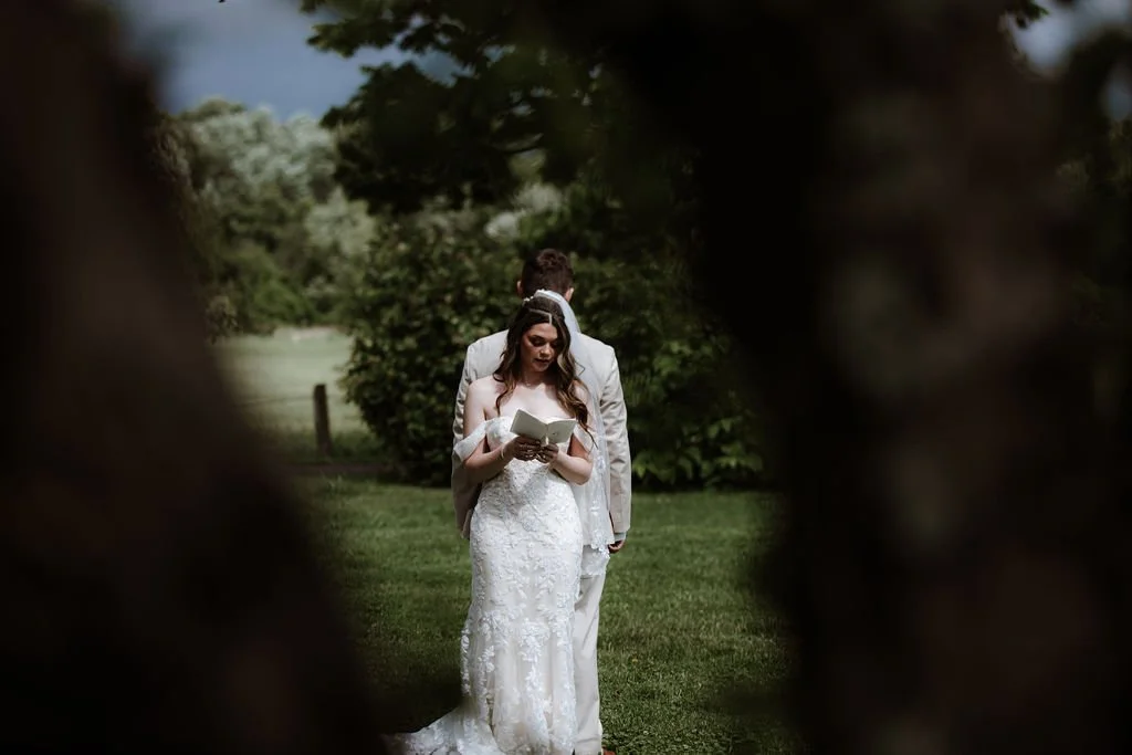 Bride in a white wedding dress reading a book, with a man in a gray suit behind her, outdoors surrounded by trees and greenery.