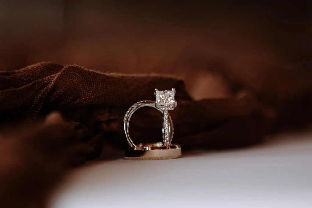 Close-up of a diamond engagement ring and wedding band displayed on a brown fabric surface.
