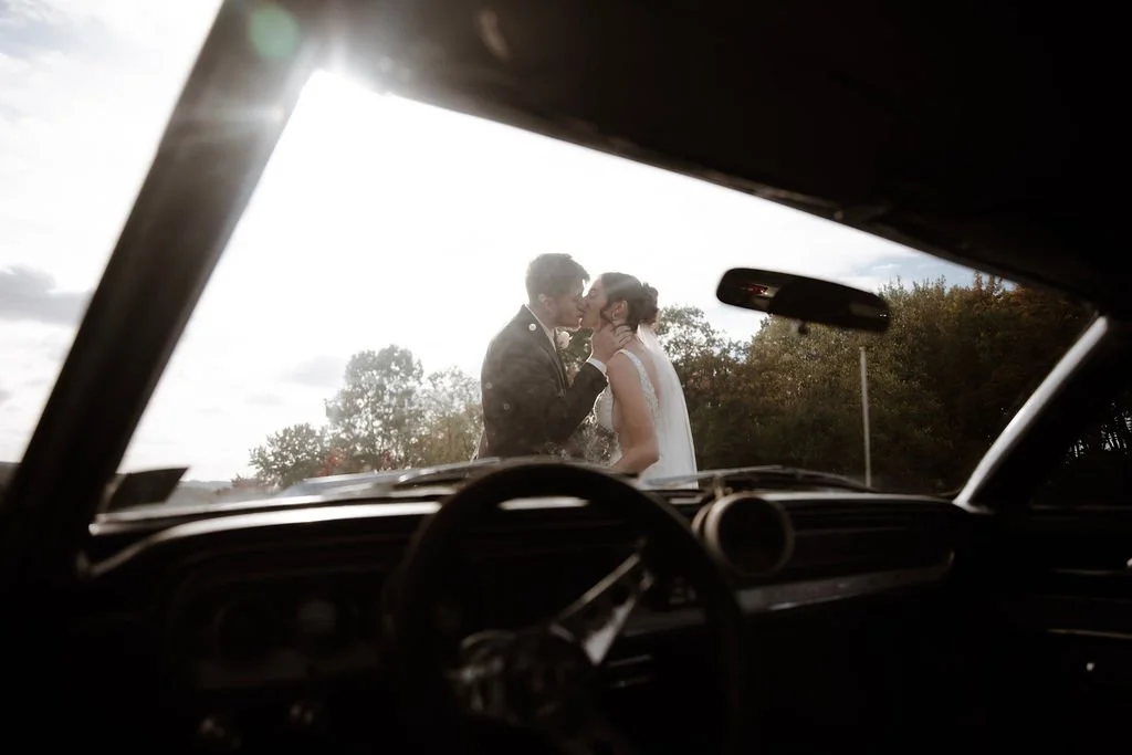 A newlywed couple kissing outdoors, seen from inside a vintage car with the dashboard and steering wheel visible in the foreground.