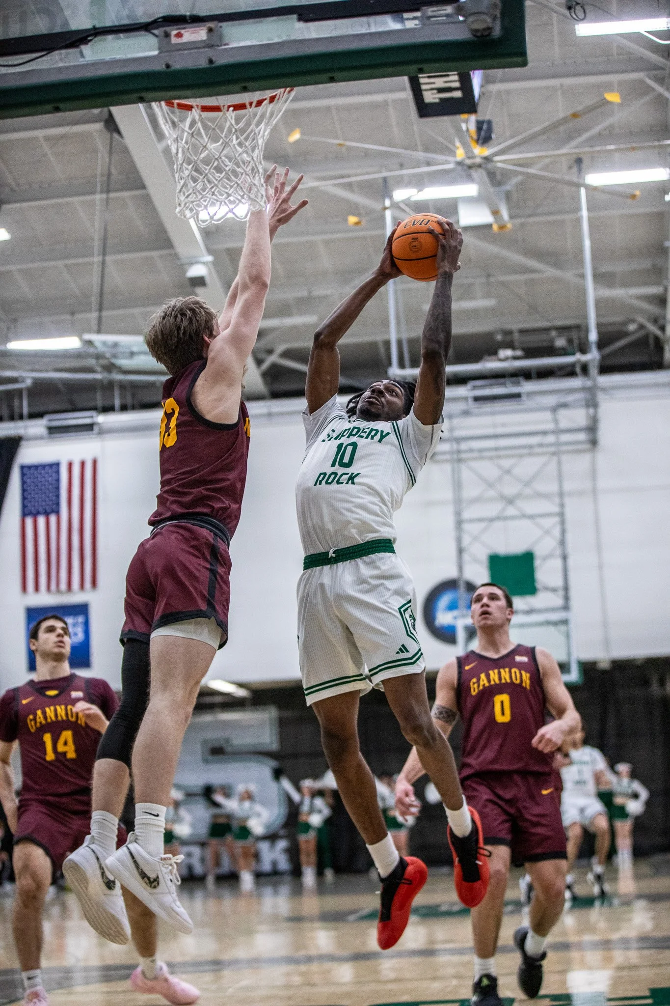 A basketball game with players in action. One player in a white jersey is jumping to make a shot while an opposing player in a maroon jersey is jumping to block him. Other players are on the court, and an American flag is visible in the background.