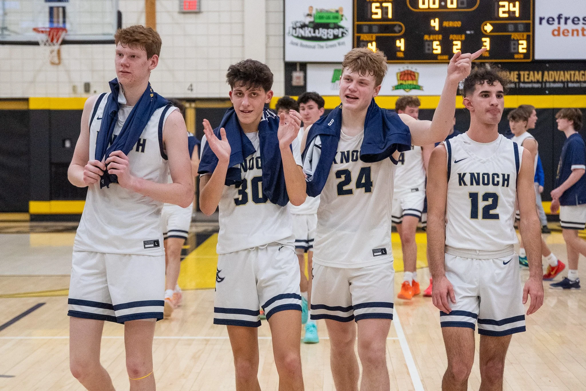 Group of young male basketball players celebrating on a gym court after a game, wearing white jerseys with blue accents and shorts. The scoreboard in the background shows the final score of 57-24.