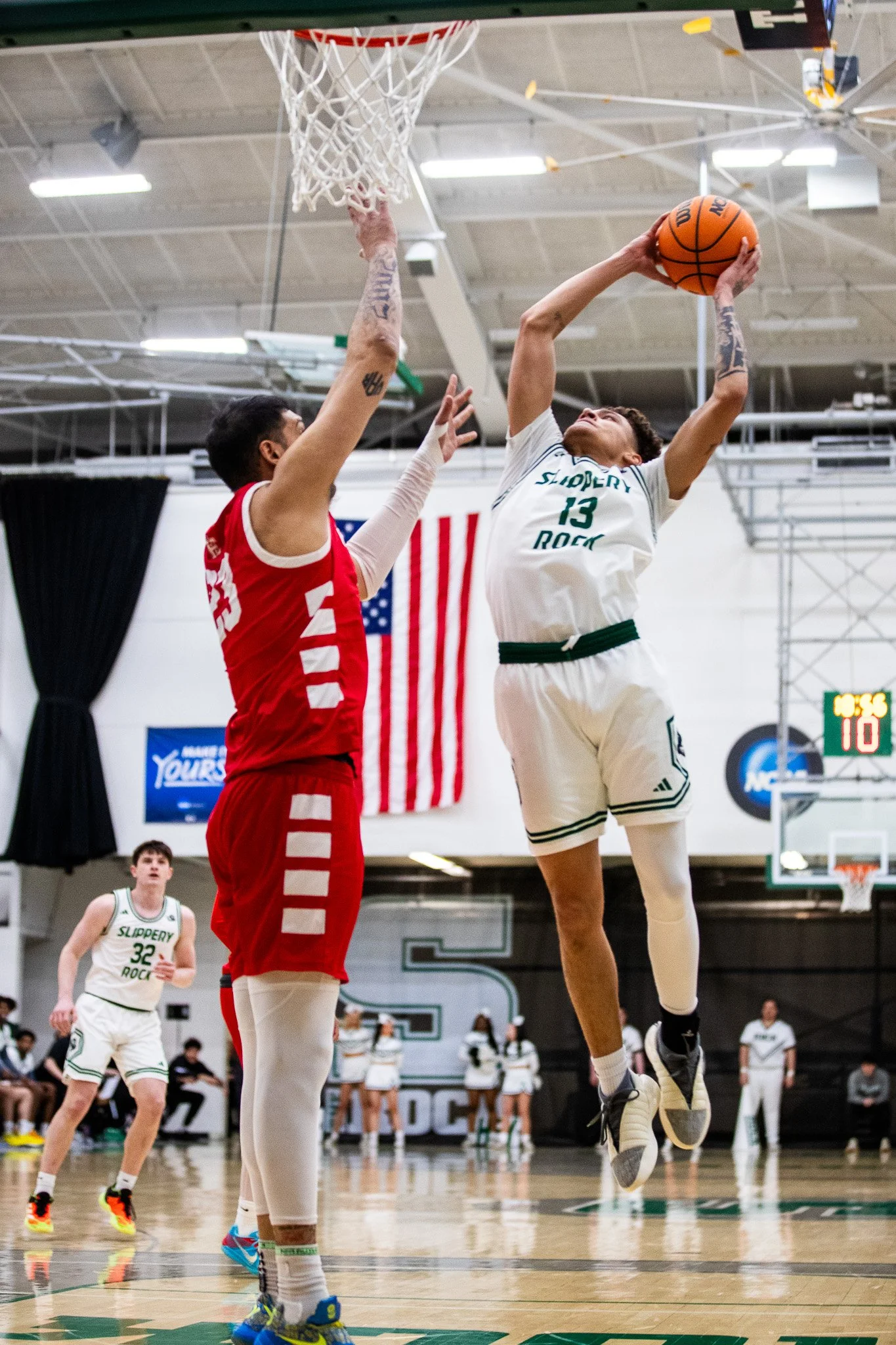 A basketball player in a white jersey jumping to shoot the ball while a player in a red jersey attempts to block the shot during a game in an indoor basketball court.