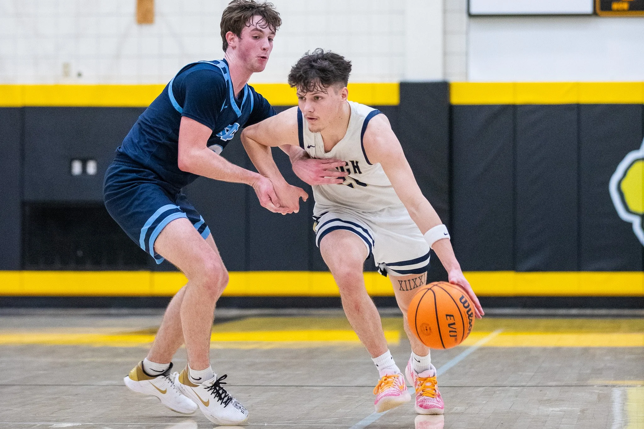 Two young men playing basketball indoors. One in a navy sports uniform is defending, and the other in a white uniform is dribbling the ball.