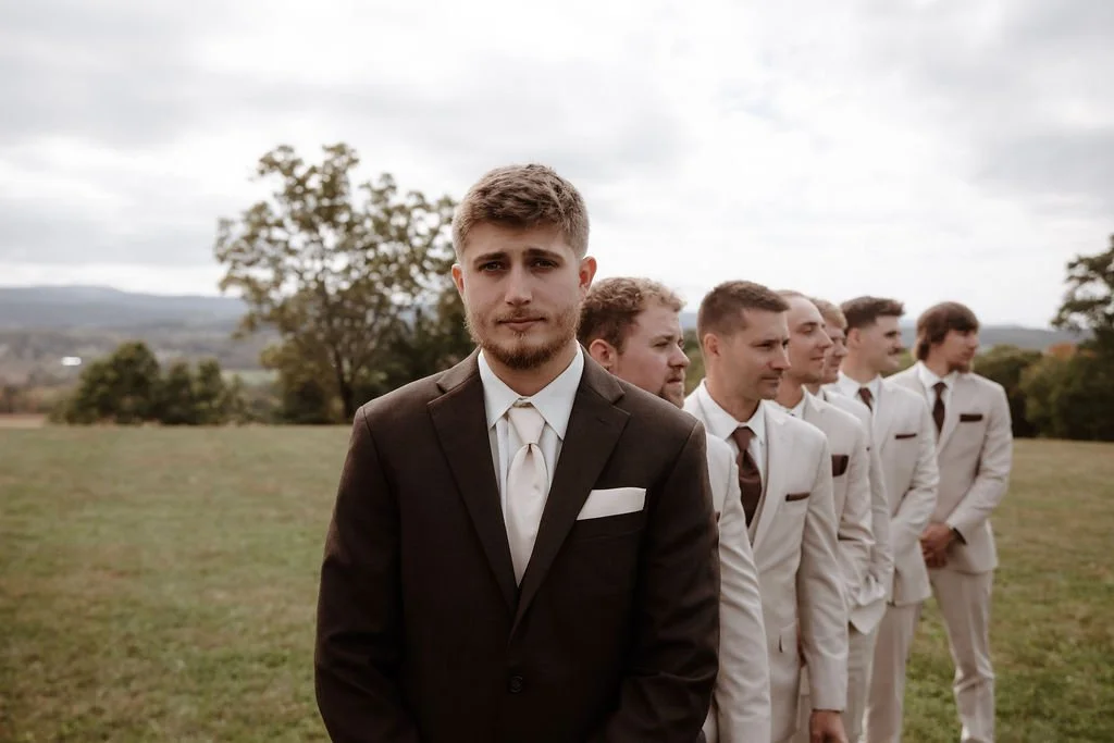 A groom and six groomsmen dressed in suits standing outdoors on a grassy field with trees and mountains in the background.