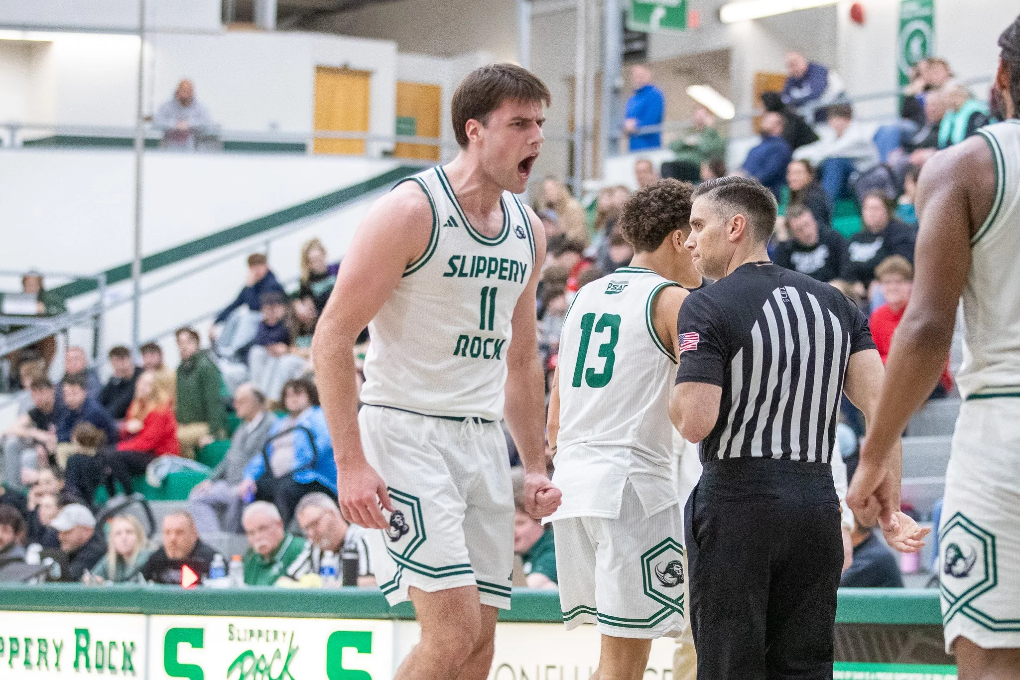 A basketball player wearing a white jersey with the number 11 and the words 'Slippery Rock' shouting at the referee during a game. Another player with number 13 is visible, and a referee is showing a card. Spectators are in the background.