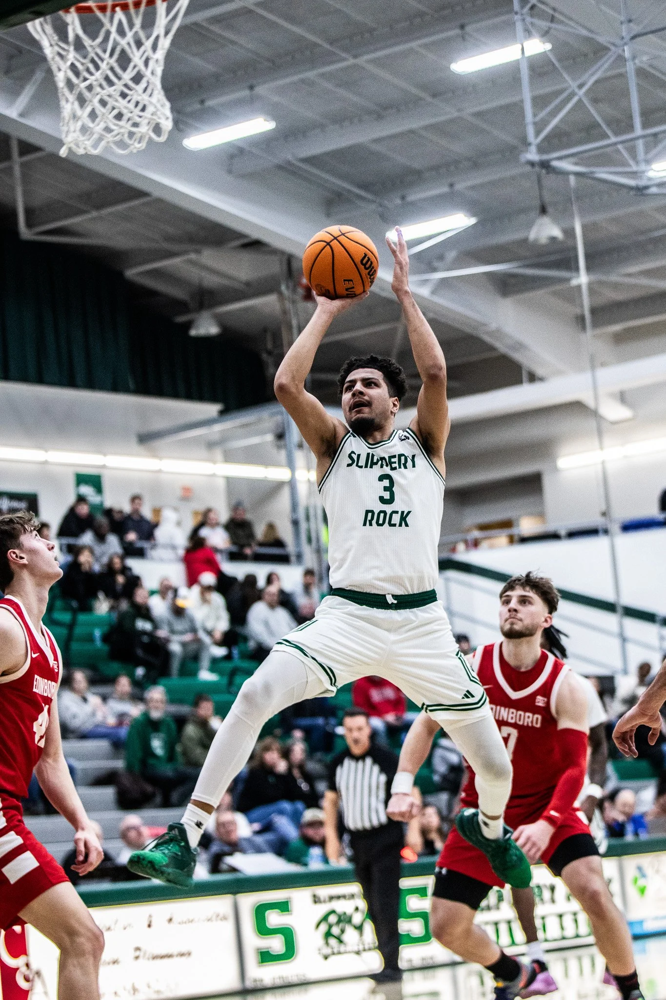 A basketball player in a white uniform with 'SLIPPERY ROCK' and the number 3, jumping and preparing to shoot or dunk the ball during a game, with opposing players in red uniforms and spectators in the background.