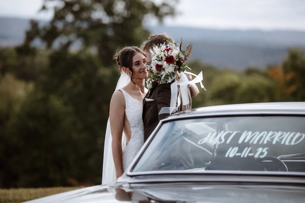 Bride and groom sharing a kiss behind a decorated car with "Just Married" and the wedding date written on the windshield, outdoor setting with trees in the background.