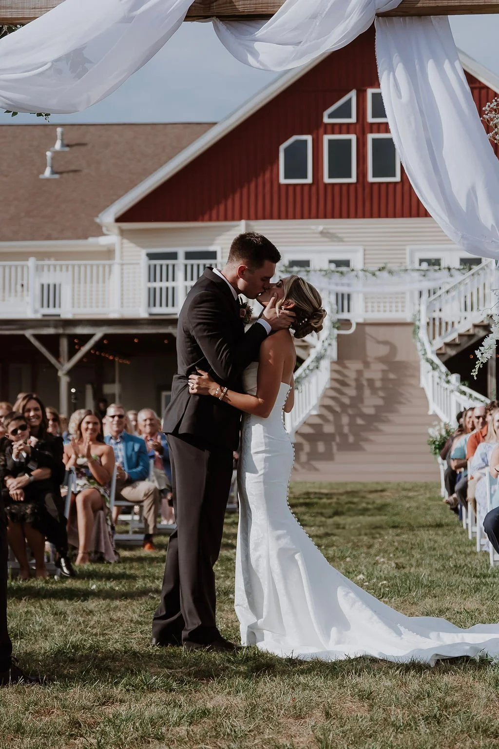 A bride and groom share a kiss during their outdoor wedding ceremony at a rustic venue with white drapery and an audience of guests sitting on either side.