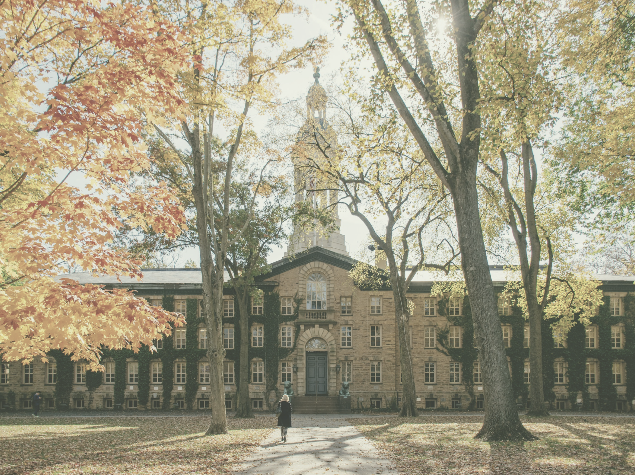 A woman walking in front of an old brick building at Princeton University, surrounded by trees with yellow and orange leaves, indicating autumn.