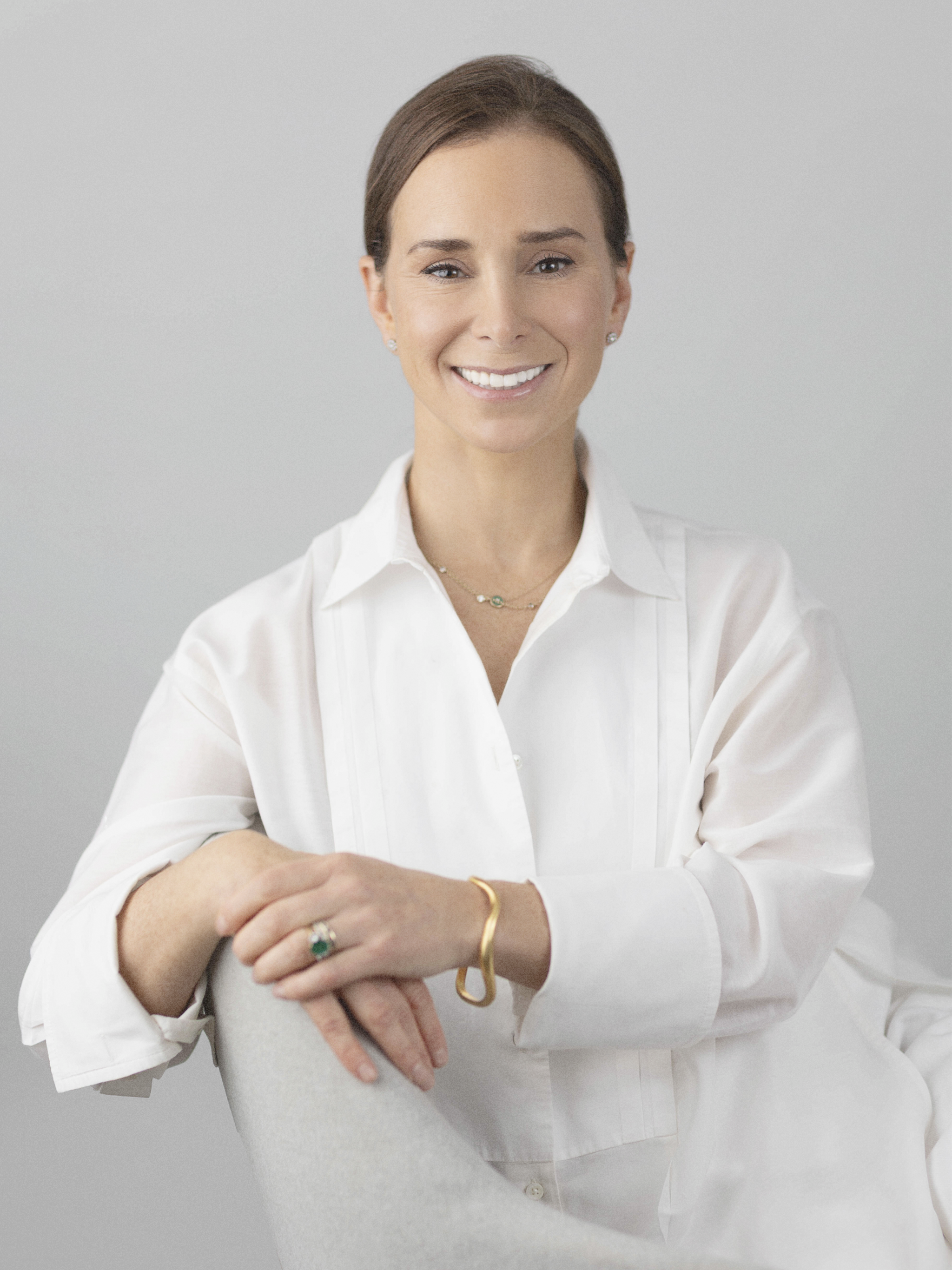 A picture of Zan Morley, wearing a white blouse, and jewelry, smiling at the camera against a plain gray background.