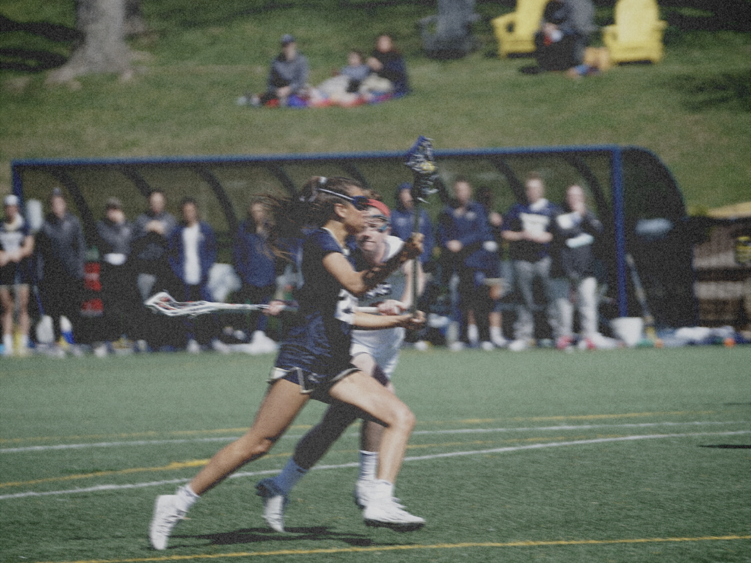 A girl running on an outdoor sports field during a lacrosse game, with spectators and other players in the background.