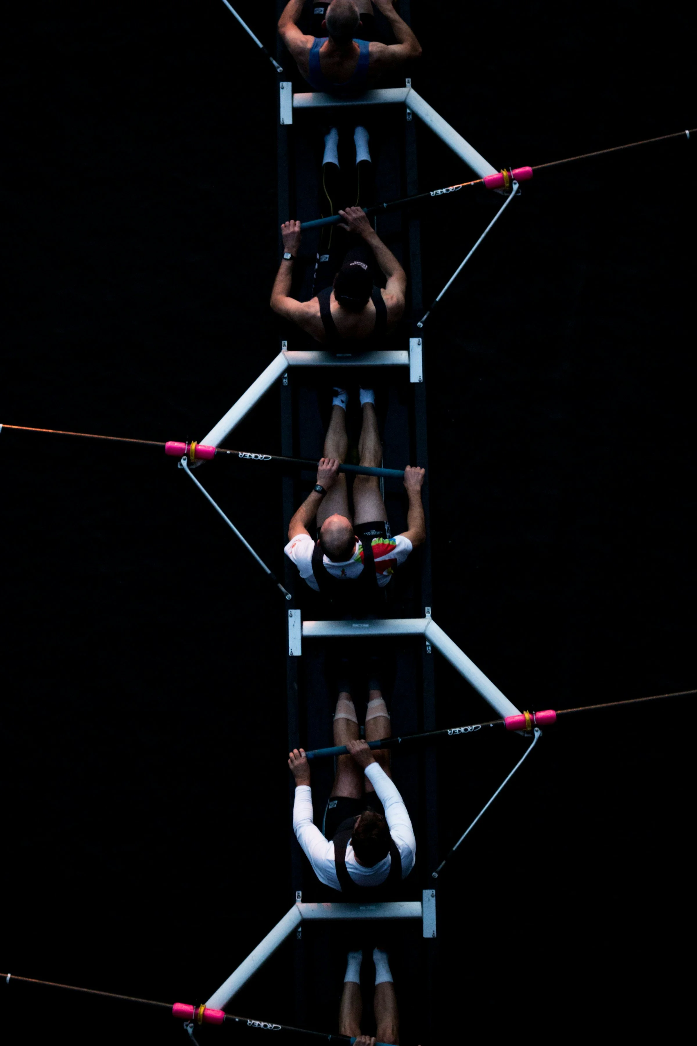 Aerial view of four rowers in a racing shell on a dark water surface, preparing for or during a rowing race.