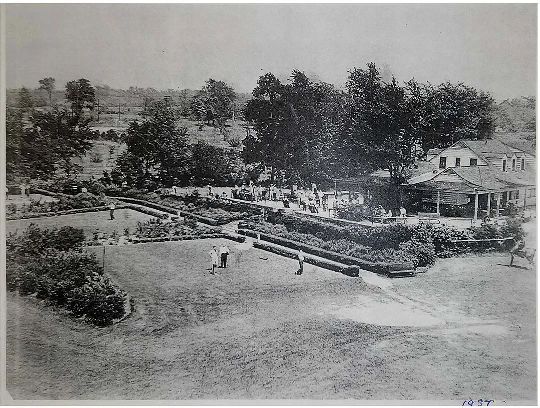 A black and white photograph of a park with people playing golf, walking, and sitting. There are trees, bushes, a building with a porch, and a fenced-in area in the background.