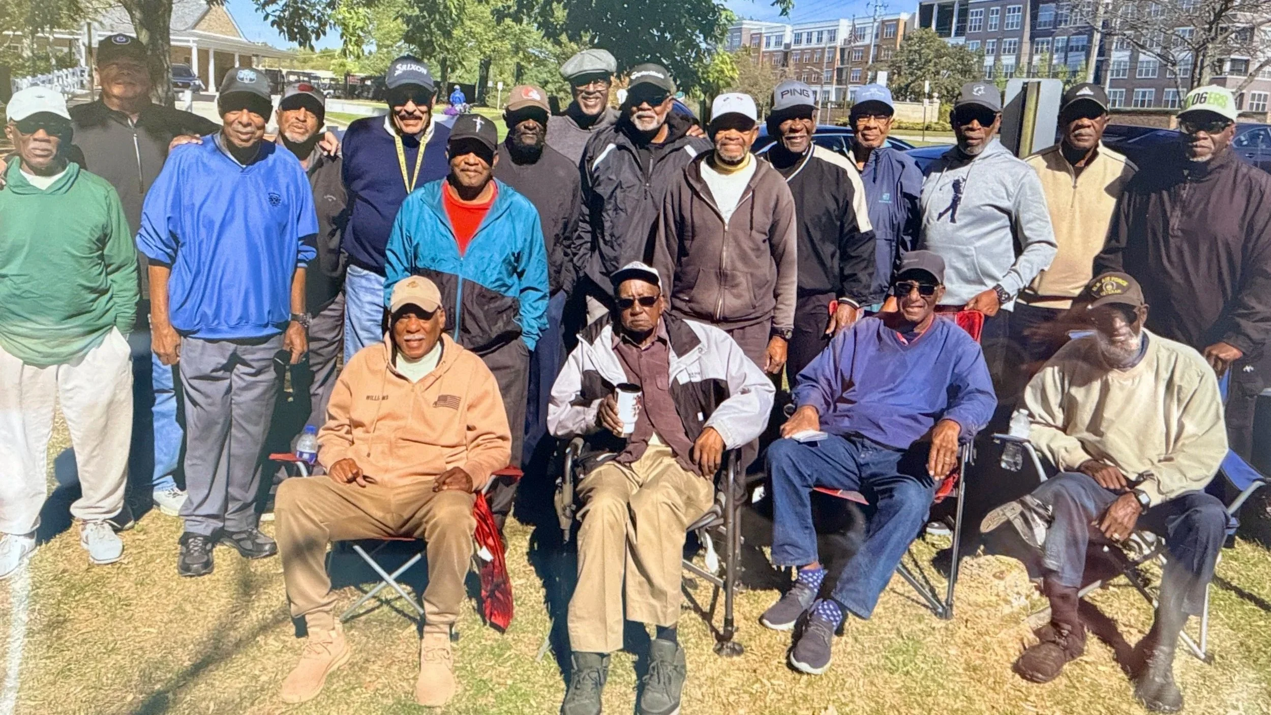 A group of men, mostly seniors, gathered outdoors in a park on a sunny day. Some men are seated, and some are standing behind them, all smiling or looking at the camera. They are dressed casually, wearing jackets, hats, and sunglasses, with a background of trees and buildings.
