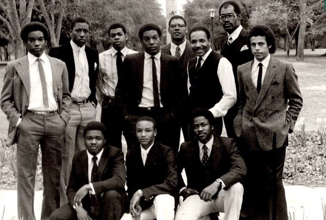 A group of eleven young men in formal suits and ties, outdoors in a park-like setting, posing for a photo.