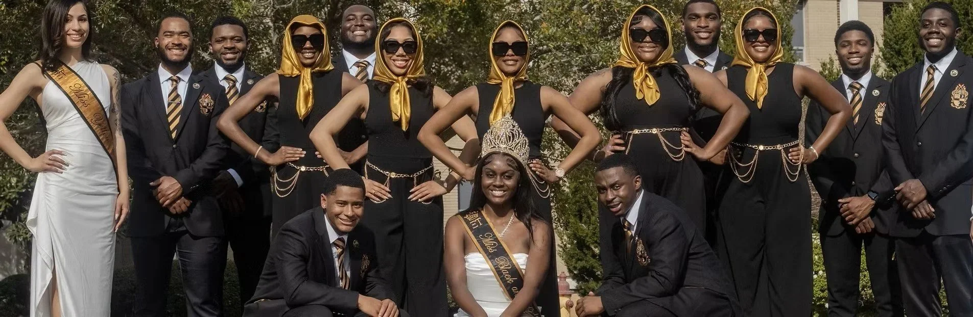 Group of young adults at a beauty pageant, with the woman in the center wearing a tiara and sash reading 'Miss Black,' and the woman on the left in a white gown with a sash reading 'Mrs. Black.' The group is dressed in black suits and dresses, some with gold accessories and headscarves, and they are posing outdoors with trees and buildings in the background.
