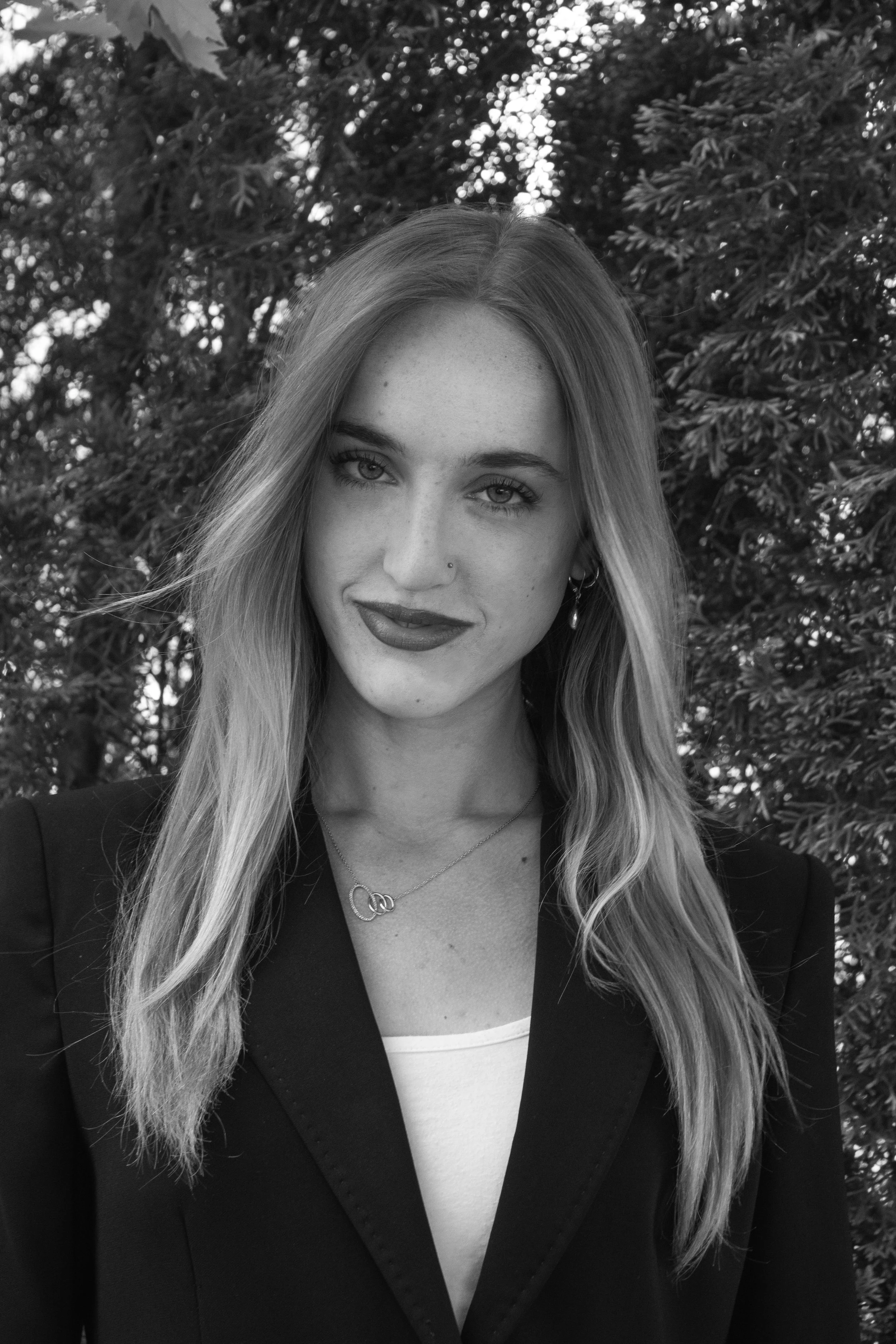 A young woman with long hair, wearing a blazer and jewelry, standing outdoors with trees in the background.
