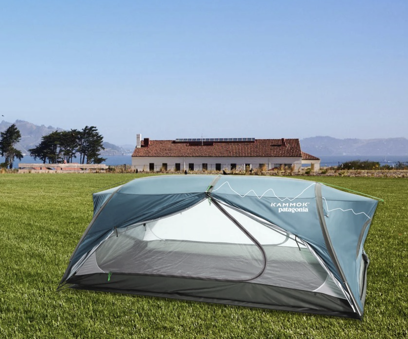 A tent set up on a grassy field with a house and trees in the background under a clear blue sky.