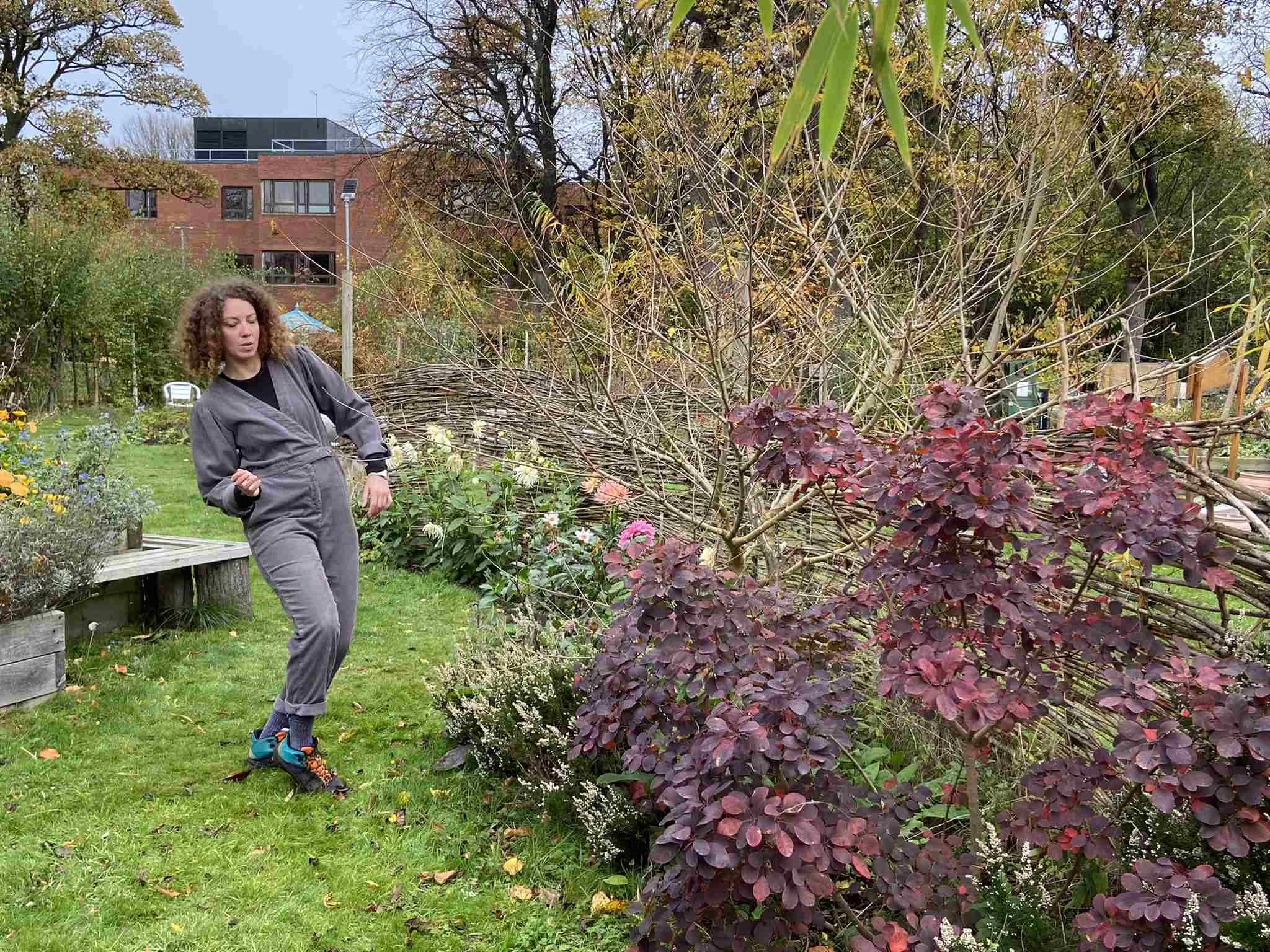 Dancer in a grey jumpsuit moves in an autumn garden beside purple-leaved shrubs and a wattle fence in the Garden Series.